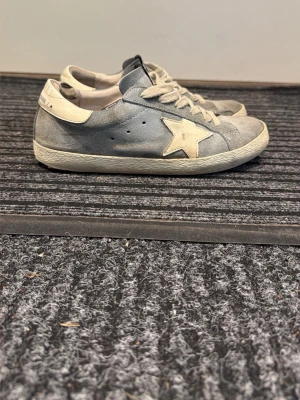 Blå sneakers med vit stjärna - Snygga blå sneakers med vit stjärndetalj på sidan och vita skosnören. Skorna har en låg profil och vit sula. Materialet ser ut att vara mocka och textil, med GGDB-logga på plösen och hälen. Perfekta för en avslappnad streetstyle-look.