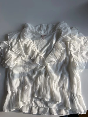 Vit blus med volanger och kort ärm - Säljer en romantisk vit blus med volanger och puffärm. Blusen har v-ringning, knappar framtill och fina detaljer med volanger längs krage, ärmar och nederkant. Perfekt till både vardag och fest! 