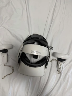Vit VR-glasögon med handkontroller - Säljer ett par vita VR-glasögon med matchande handkontroller. Glasögonen har en modern, rundad design med justerbart huvudband och mjuka kuddar för komfort. Materialet är plast med detaljer i svart och vitt. Perfekt för gaming och VR-upplevelser.
