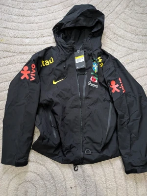 Svart Brasilien vindjacka Nike - Snygg svart vindjacka från Nike med Brasilien-tema. Jackan har huva, dragkedja framtill och flera loggor i gult, rött och grönt. Märken som 'vivo', 'itau' och Brasil-logga syns tydligt. Perfekt för dig som gillar sportig stil och streetwear.