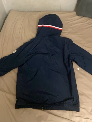 Mörkblå Moncler vindjacka med huva - Snygg mörkblå vindjacka från Moncler med huva och dragkedja. Jackan har en vit och röd rand på huvan samt Moncler-logga på ärmen. Tillverkad i lätt och tåligt material, perfekt för blåsiga dagar. Klassisk passform och elastisk nederkant.
