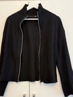 Ribbad cardigan med dragkedja - En svart ribbstickad cardiganjacka med hög krage och silverfärgad dragkedja. Jackan har långa ärmar och är gjord i ett mjukt, tjockt material som känns riktigt cozy. Perfekt att slänga på sig när det är kyligt ute.