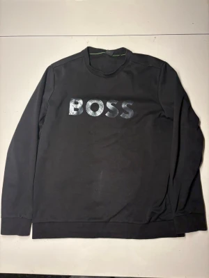 Svart sweatshirt från BOSS - Snygg svart sweatshirt från BOSS med stor, glansig logga på bröstet. Tröjan har rund halsringning och långa ärmar. Tillverkad i mjukt material som känns skönt mot huden. Perfekt för en clean och stilren look.