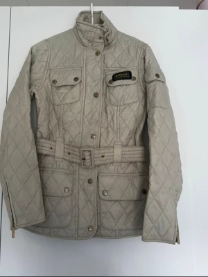 Beige quiltad jacka från Barbour - Snygg beige quiltad jacka från Barbour med bälte i midjan, hög krage och flera fickor framtill. Jackan har guldfärgade knappar, Barbour International-märke på bröstet och lång ärm. Perfekt för dig som gillar klassisk och stilren look.