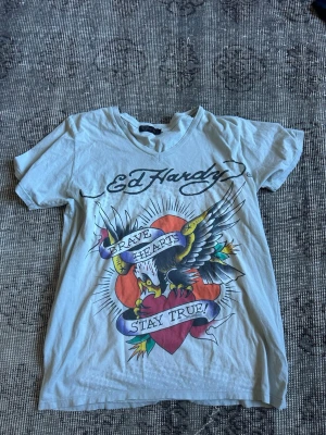 Ed Hardy t-shirt med örn och hjärta - Snygg vit Ed Hardy t-shirt med färgglatt tryck av en örn och hjärta på framsidan samt svartvitt tryck på baksidan. Texten 'Brave Hearts Stay True' och klassisk Ed Hardy-logga. T-shirten har korta ärmar och v-ringad hals. Perfekt för dig som gillar statement-plagg.