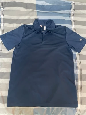 Adidas Golfpike  - Snygg mörkblå pikétröja med korta ärmar och klassisk krage. Tröjan har knappslå framtill och diskret vitt tryck på ena ärmen. Tillverkad i ett funktionsmaterial som passar perfekt till träning eller sport.
