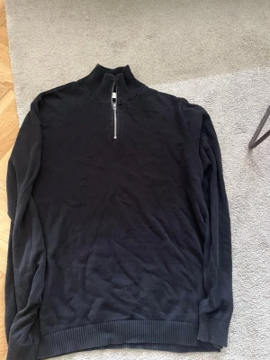 Svart tröja med halv dragkedja - En svart långärmad tröja med halv dragkedja framtill och ribbade muddar vid ärmslut och nederkant. Tröjan har en hög krage och är tillverkad i ett mjukt material som passar perfekt för lager på lager-stil.