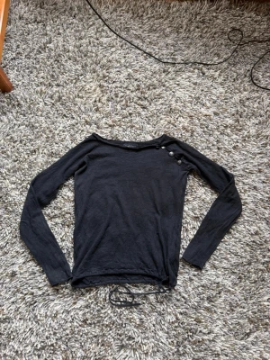 Svart långärmad topp Zadig & Voltaire - Svart långärmad topp från Zadig & Voltaire med rund halsringning och dekorativa knappar på ena axeln. Tunn och mjuk kvalitet, perfekt för lager på lager. Dragsko i nederkant för justerbar passform. Storlek M men sitter som en XS. Material: Silke och kashmir.
