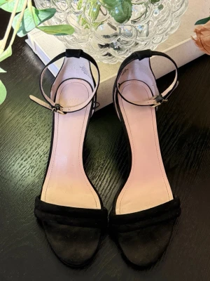 Svarta pumps med ankelrem - Stilrena svarta pumps med öppen tå och smal ankelrem. Skorna har en klassisk klack och är tillverkade i mockaliknande material. Perfekta för dig som gillar en enkel men elegant look.