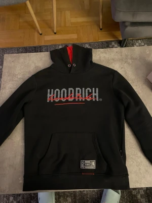 Svart hoodie från Hoodrich - Snygg svart hoodie från Hoodrich med stor logga i grått och rött framtill. Hoodien har huva med röd insida, dragsko och en stor magficka. Perfekt för en streetwear-look och tillverkad i mjukt material för extra komfort.