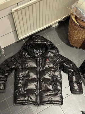 Svart glansig pufferjacka med huva - Säljer en svart, glansig pufferjacka med huva och dragkedja framtill. Jackan har två stora fickor på framsidan och en liten röd logga på bröstet. Perfekt för kalla vinterdagar och riktigt skön att bära. Materialet är syntet och jackan är vadderad för extra värme.