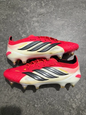 Adidas Predator fotbollsskor röd/vit - Säljer ett par Adidas Predator fotbollsskor med snygg design i rött, vitt och svart. Skorna har metallskruvdobbar och en låg profil för bästa grepp på planen. Ovandelen är i syntetmaterial och har klassiska Predator-detaljer på sidorna. använd i 1 måndag och priset kan deskuteras
