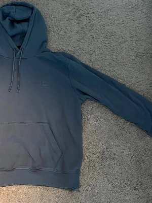 Blå hoodie från boss - Säljer en stilren blå hoodie med huva och dragsnören. Hoodien har en stor magficka framtill och ett diskret broderat märke på bröstet. Tillverkad i mjukt bomullsmaterial och har ribbade muddar vid ärmslut och nederkant. Perfekt för en avslappnad look. Säljer den för 250 kr kan gå med på priser till 170 kr! Den är medium i storlek men än väldigt liten så passar nästan bara på small ändå!