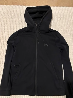 Svart hoodie med dragkedja från BOSS - Svart hoodie från BOSS med hel dragkedja framtill, två fickor med dragkedja och diskret BOSS-logga på bröstet. Klassisk passform med huva och långa ärmar. Perfekt för en stilren och avslappnad look.