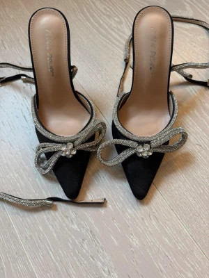 Svarta pumps med strass och rosett - Säljer ett par svarta pumps med spetsig tå och smal klack i metall. Skorna har dekorativa strassband som bildar en stor rosett framtill och remmar med strassdetaljer. Perfekta för dig som vill sticka ut med extra bling. Använd fåtal gånger