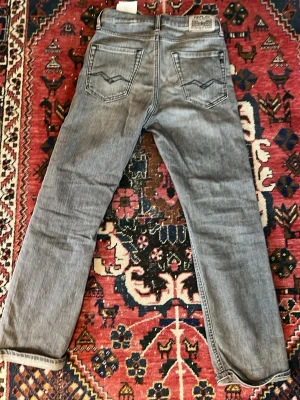 Grå raka jeans från Replay - Säljer ett par grå jeans från Replay med raka ben och klassiska bakfickor med snygga sömmar. Jeansen har en avslappnad passform och är tillverkade i ett mjukt denimtyg. Perfekta för en chill och stilren look.