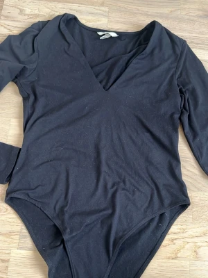 Svart långärmad body med V-ringning - En enkel och stilren svart body med lång ärm och djup V-ringning. Tillverkad i mjukt stretchigt tyg som sitter skönt mot kroppen. Perfekt att matcha med jeans eller kjol för en clean look.