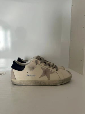Golden goose skor  - Snygga vita sneakers med grå stjärna på sidan och mörkblå häl. Skorna har grå snören och en något smutsad sula för en trendig, använd look. Märkningen GGDB/STAR syns på sidan. Perfekta för dig som gillar streetstyle och unika detaljer.