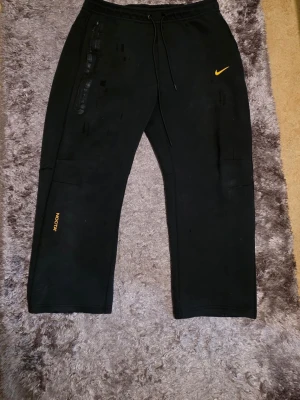 Svarta Nike mjukisbyxor med tryck - Svarta mjukisbyxor från Nike med gul logga på vänster lår och texten NOCTA på höger ben. Byxorna har dragsko i midjan, raka ben och flera fickor med dragkedja. Perfekta för en avslappnad och sportig stil. Dem är defekter därför sälja för billigt