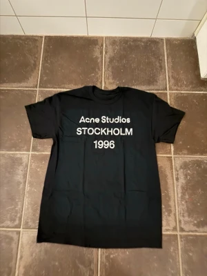 Svart Acne Studios t-shirt - Svart t-shirt från Acne Studios med vit text 'Acne Studios STOCKHOLM 1996' på bröstet. Klassisk passform med rund hals och korta ärmar. Perfekt för dig som gillar stilrena och trendiga plagg. Acne är lite oversize men passar som en vanlig S