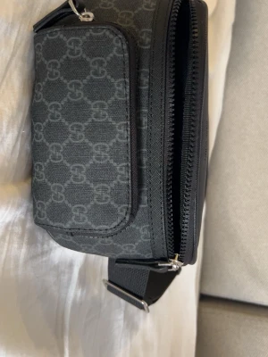 Svart handväska från Gucci - Snygg svart handväska från Gucci med klassiskt GG-mönster i grått. Väskan har en justerbar axelrem, två fack med dragkedja och är tillverkad i slitstarkt tygmaterial. Perfekt för dig som gillar stilrena accessoarer med lyxig känsla.