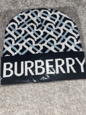Burberry monogrammönstrad mössa - Snygg mössa från Burberry med svart, vit och ljusblått monogrammönster och stor logotyp framtill. Mössan har uppvikt kant och är stickad i mjukt material som håller dig varm. Perfekt accessoar för att lyfta din outfit med en lyxig touch.