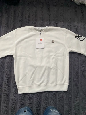 Vit sweatshirt från Moncler - Snygg vit sweatshirt från Moncler med rund halsringning och ribbade muddar. Tröjan har Moncler-logga broderad på bröstet och en svart detalj på ena ärmen. Perfekt för en clean och stilren look. Tröjan är helt ny och aldrig använd! Storleken är inte helt rätt så den är egenlitgen S och passar folk mellan 170-185. Man får tröjan i en moncler påse 