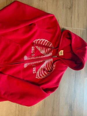 One of one hoodie - Snygg röd hoodie från One of One med dragkedja och huva. Framsidan har ett coolt skelettmotiv i Rhinestones. Tillverkad i mjukt material som känns skönt mot huden. Perfekt för dig som vill sticka ut med en unik stil. Köpt av mig limiterad sen långt tillbaka för 1.6 grymt skick inte en ända sten har lossnat!!