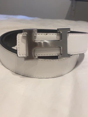 Vitt skärp med stor H-spänne - Snyggt vitt skärp med ett stort, blankt H-format metallspänne. Skärpet har stilrena sömmar och är gjort i slätt skinn. Perfekt accessoar för att lyfta din outfit med en clean och modern look.