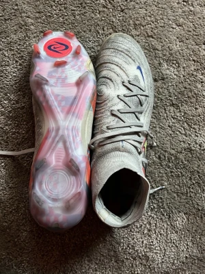 Nike fotbollsskor  - Säljer ett par gråa fotbollsskor med högt skaft och snörning. Skorna har ett vågigt mönster på ovandelen och färgdetaljer i blått och orange. Sulan är genomskinlig med rosa och röda inslag. Perfekta för dig som vill sticka ut på planen.