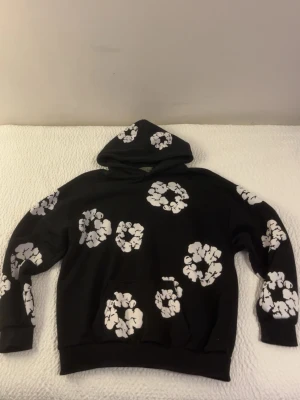 Denim tears hoodie svart vit  - Säljer en svart hoodie med stora vita blommönster över hela tröjan. Hoodien har en klassisk huva och magficka framtill. Mjuk och skön passform, perfekt för dig som gillar streetwear och vill sticka ut lite extra.