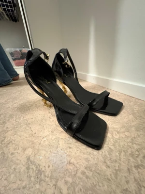 Svarta klackar från YSL - Snygga svarta klackar från YSL med fyrkantig tå och tunna remmar. Klacken är i guld med YSL-logga, vilket ger en lyxig touch. Skorna har elegant siluett som sticker ut.