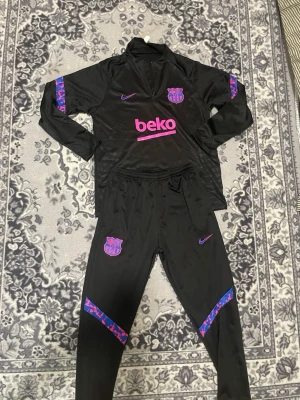 Svart FC Barcelona träningsset Nike - Säljer ett svart träningsset från Nike och FC Barcelona med lila och rosa detaljer. Setet består av långärmad tröja med halv dragkedja och matchande byxor med klubbmärke och Nike-logga. Materialet är troligtvis polyester, perfekt för träning eller uppvärmning.