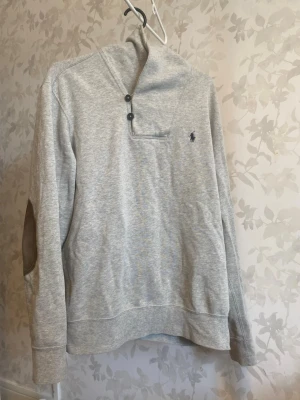  Polo Ralph Lauren med en klassisk design.  -  Polo Ralph Lauren med en klassisk design. Ljusgrå sweatshirt med hög krage och tre knappar vid halsen. Tröjan har ett diskret broderat logotyp på bröstet och mockadetaljer på armbågarna. Tillverkad i mjukt material och har långa ärmar med breda muddar.