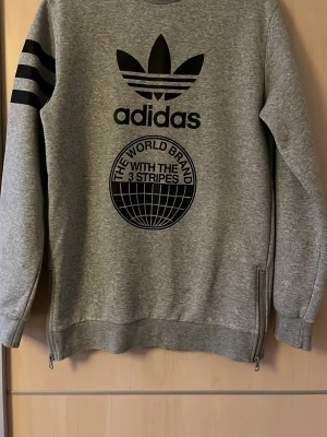 Grå Adidas hoodie med tryck - Snygg grå hoodie från Adidas med stort svart tryck framtill och klassiska tre ränder på ena ärmen. Tröjan har rund hals, muddar vid ärmslut och nederkant samt dragkedjor i sidorna. Tillverkad i mjukt sweatshirtmaterial, perfekt för en avslappnad stil.