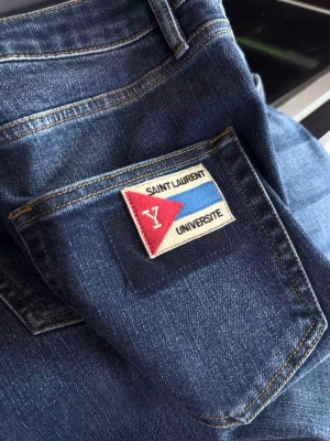 Blå jeans Saint Laurent Université - Snygga blå jeans från Saint Laurent med patch på bakfickan där det står 'Université'. Klassisk femficksmodell med guldfärgade nitar och knapp. Jeansen har en rak passform och är gjorda i denim med tydliga sömmar.