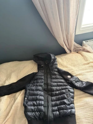 Svart moncler  pufferjacka - Säljer en svart moncler pufferjacka med huva och ribbade muddar. Jackan har quiltad framsida och ärmar i mjukare material, dragkedja framtill och två fickor med knappstängning. Perfekt för kalla vinterdagar och riktigt snygg streetstil.