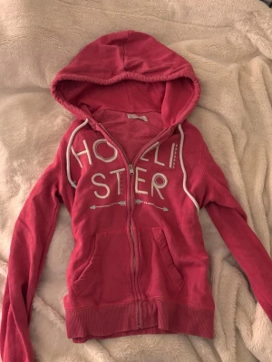 Rosa hoodie med dragkedja från Hollister vintage  - Säljer en rosa hoodie från Hollister med vit text och pilmotiv på bröstet. Hoodien har dragkedja framtill, två fickor och vit dragsko i huvan. Tillverkad i mjukt bomullsmaterial och har ribbade muddar vid ärmslut och nederkant. Stl M men liten i storlek!!