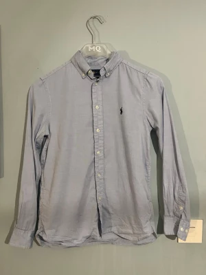 Ljusblå skjorta från Ralph Lauren - Klassisk ljusblå skjorta från Ralph Lauren med button-down krage och broderad logga på bröstet. Skjortan har långa ärmar och knäppning framtill. Perfekt för en stilren och avslappnad look.