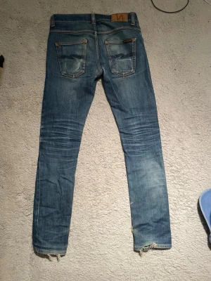 Blå slim/ raka  jeans från Nudie Jeans - Säljer ett par blå slim/ raka   jeans från Nudie Jeans med slitningar och ljusa tvättade partier. Jeansen har klassiska bakfickor med Nudie-söm, normal midja och råa benslut. Perfekta för en avslappnad och trendig look. Bra fades