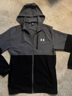 Under armour jacka - Snygg tvåfärgad vindjacka från Under Armour i grått och svart. Jackan har huva, hel dragkedja framtill och en bröstficka med dragkedja. Perfekt för dig som vill ha en sportig look och gillar funktionella detaljer. Strlk L