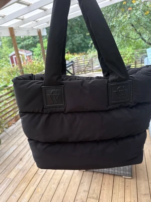 Svart vadderad totebag - Snygg svart shoppingväska i puffad design med breda axelband och flera innerfack, inklusive ett med dragkedja. Väskan har en modern look och är tillverkad i vadderat tyg som ger en mjuk känsla. Perfekt för dig som vill ha plats för allt du behöver under dagen.