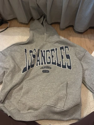 Grå glittrig hoodie med Los Angeles-tryck - Helt ny Grå glittrig hoodie med stor huva och trycket 'Los Angeles California' i mörkblått och vitt på bröstet. Hoodien har en klassisk passform, ribbade muddar och magficka. Perfekt för en chill och avslappnad stil. Materialet är mjukt och bekvämt, troligen bomullsblandning.
