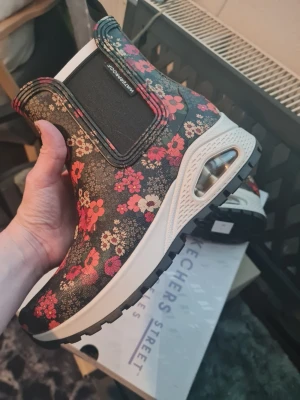 Blommiga Skechers regnstövlar med sportig sula - Snygga regnstövlar med svart bas och färgglatt blommönster i rött, rosa och beige. Skorna har elastiska paneler på sidorna och en vit, sportig sula med synlig luftbubbla. Perfekta för dig som vill sticka ut med din stil och gillar bekväma skor. Stl 37. Helt nya och oanvända i kartong. Nypris ca 950 kr.