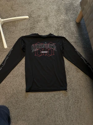 Svart långärmad träningströja Gymshark - Svart långärmad träningströja från Gymshark med tryck i vitt och rött framtill samt vita detaljer längs ärmarna. Tillverkad i ett syntetmaterial som passar perfekt för träning. Tröjan har en normal passform och är riktigt bekväm för aktiva dagar. Tröjan är storlek L men passar nog mer på storlek M