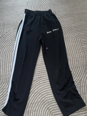 Palm Angels svarta mjukisbyxor - Svarta mjukisbyxor från Palm Angels med vit sidostripes och logga tryckt på vänster sida. Byxorna har resår i midjan och raka ben. Materialet ser ut att vara polyester eller liknande för en sportig vibe. Perfekta för en chill och trendig look.
