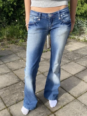 Blå bootcut jeans med låg midja - Säljer ett par blå bootcut jeans med låg midja och slitningar framtill. Jeansen har fickor med knappdetaljer och ljusare tvättade partier på låren. Perfekta för en avslappnad och trendig look. slitningar längst ner