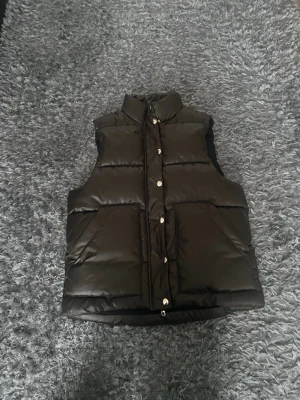 Svart puffad dunväst med hög krage - Snygg svart dunväst med puffad design och hög krage. Västen har stora fickor framtill och stängs med silverfärgade tryckknappar. Perfekt för lager på lager och ger en cool streetwear-look. Materialet är glansigt och quiltat för extra värme.