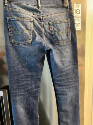 Blå Levi's jeans med raka ben - Säljer ett par klassiska blå Levi's jeans med raka ben och normal midja. Jeansen har två bakfickor med den ikoniska V-formade sömmen och en liten röd Levi's-tag på höger ficka. Tillverkade i slitstark denim med snygg tvätt och kontrastsömmar.