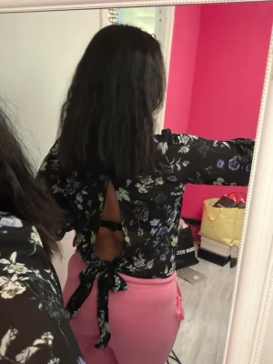 Blommig svart blus med öppen rygg - Snygg svart blus med blommönster i lila, vitt och grönt. Blusen har lång ärm och är gjord i ett tunt, transparent material. Öppen rygg med knytband nedtill ger en trendig och unik look. Perfekt för dig som vill sticka ut lite extra.